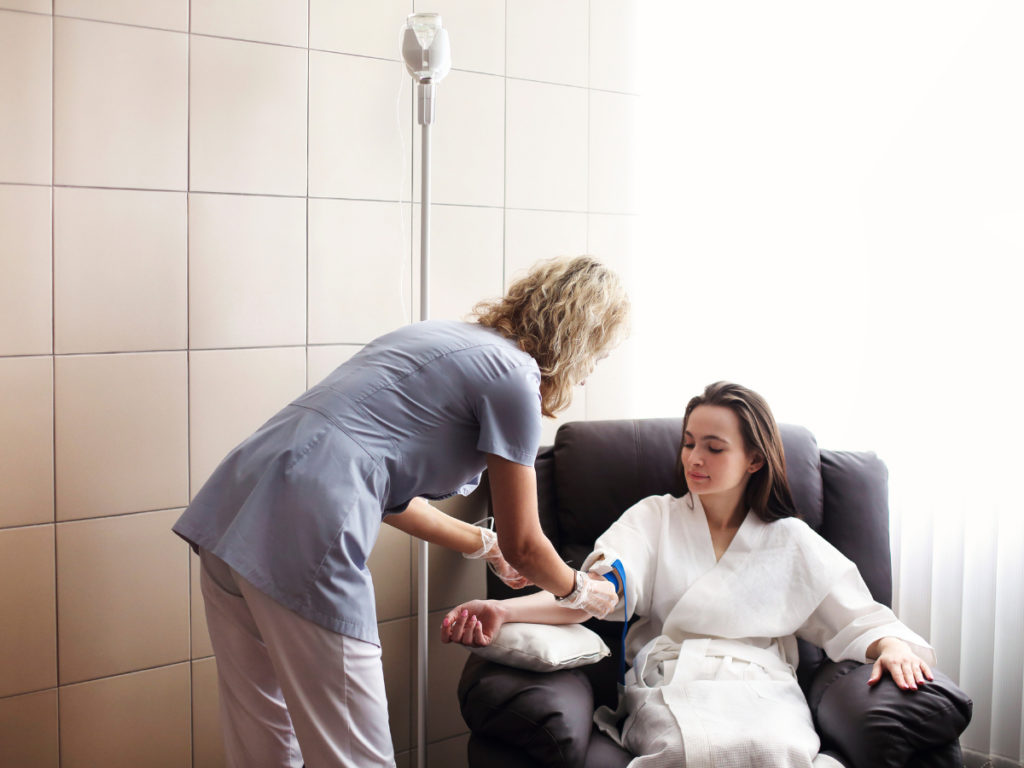 a woman in a white medical robe sitting on a plush chair receiving a medical injection from a medical professional wearing gloves.