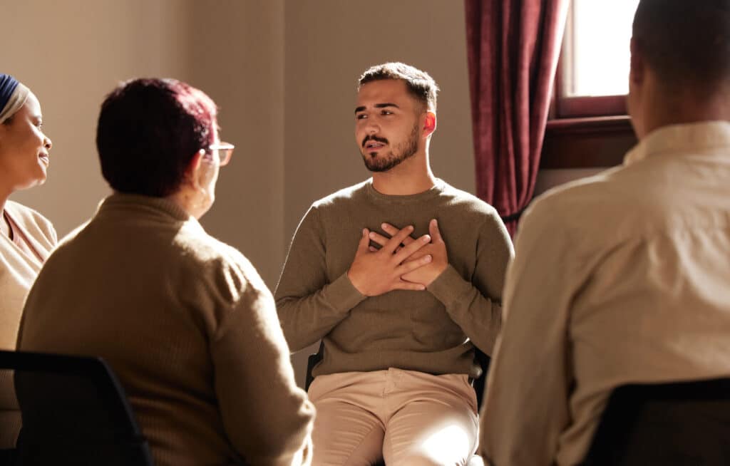A man holds his hands over his heart as he shares his experiences in recovery with an addiction recovery support group around him
